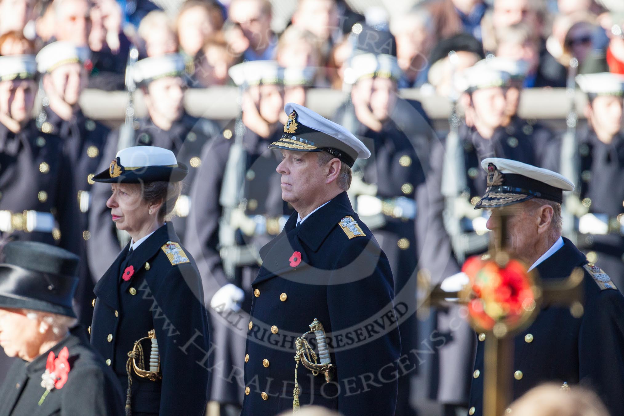 The golden cross with the poppies carried by the cross bearer, out of focus, and HRH The Princess Royal, HRH The Duke of York and HRH The Duke of Edinburgh.