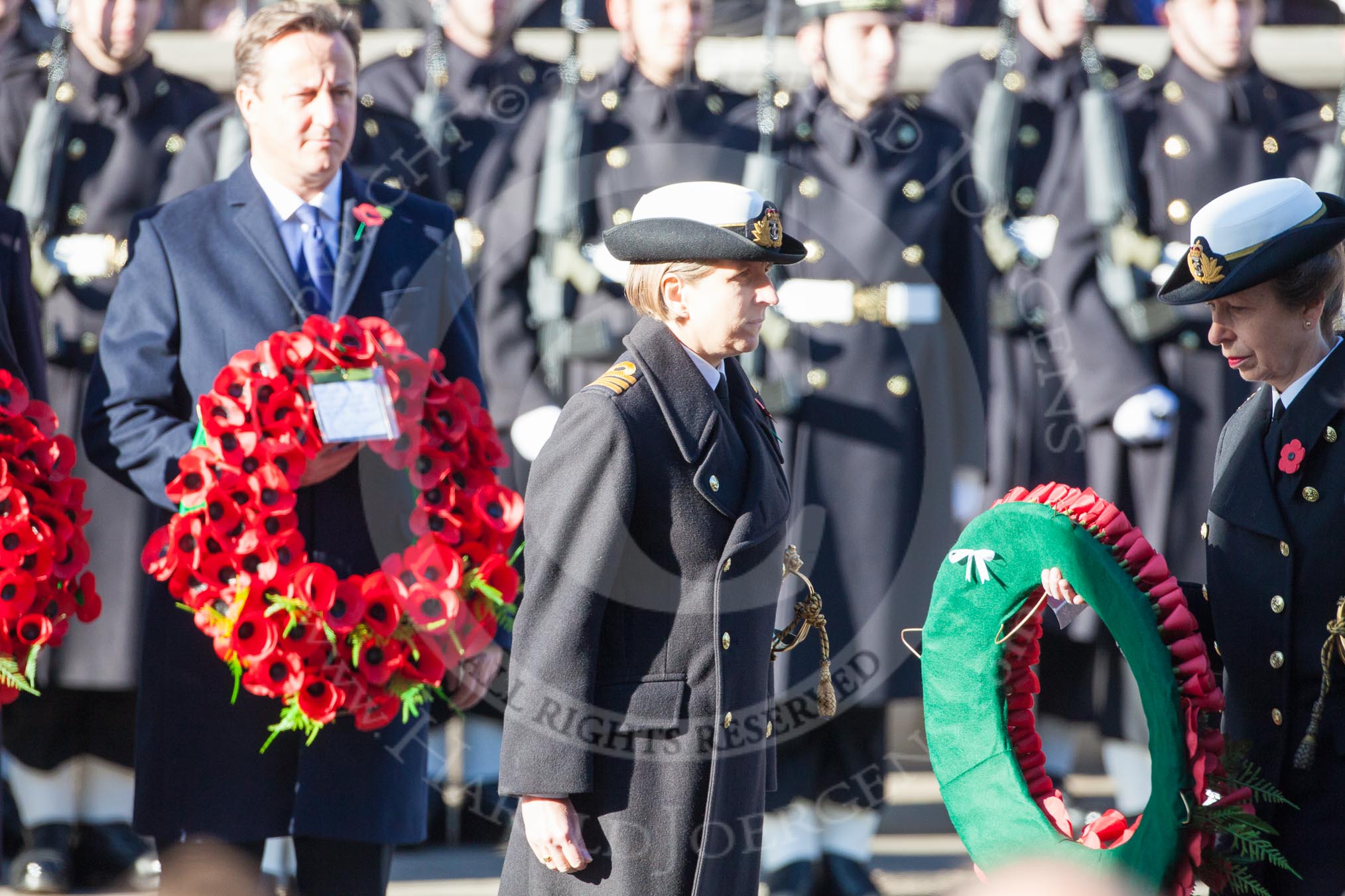 Commander Anne Sullivan, Royal Navy, handing, as Equerry, the wreath to HRH The Princess Royal. In the background the Prime Minister, David Cameron.